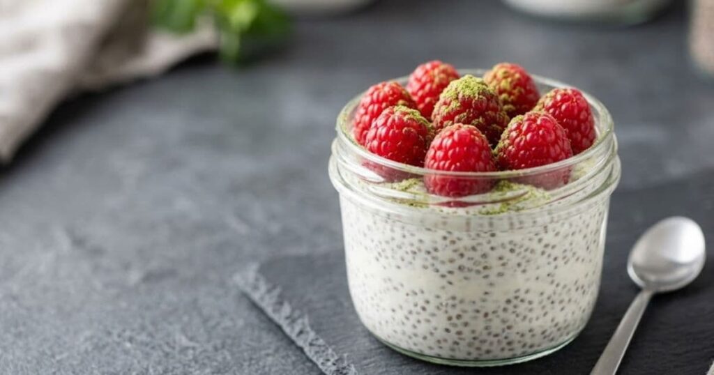 Superalimentos Funcionais - Foco em um pote de vidro com pudim de chia, framboesas frescas e pó de matcha em uma mesa de ardósia.